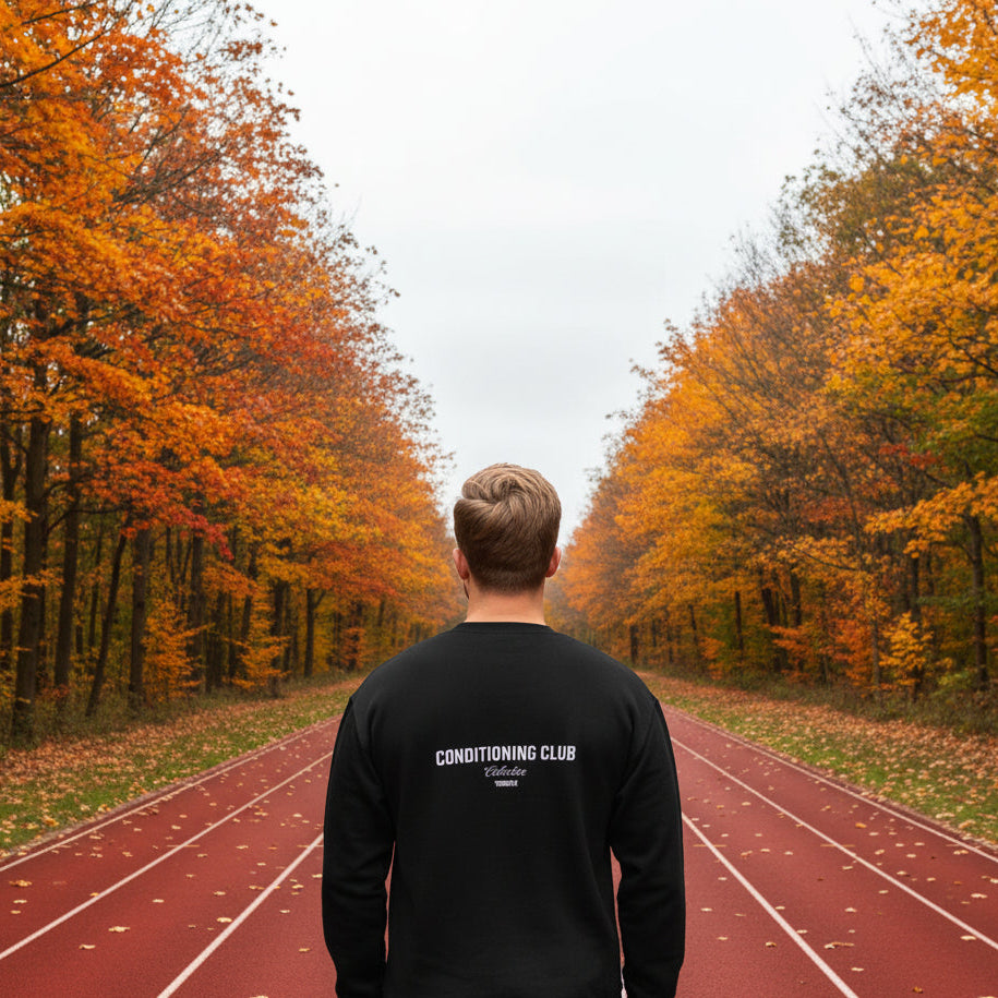 Person wearing a black sweatshirt with 'Conditioning Club Collective' text on the back against a white brick wall.